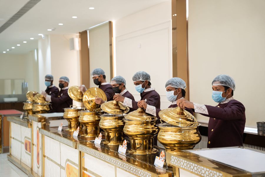 Staff in maroon uniforms lifting the lids of brass chafing dishes along a jaali-panelled buffet counter